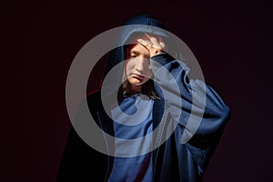 Portrait of a sad lonely teenager / young girl in a black hoodie in a black-red background. Eyes closed, hands on head. Problems