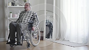 Portrait of sad old Caucasian disabled man turning wheelchair from window and rolling away. Male retiree in depression