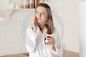 Portrait of sad pensive woman talking on the mobile phone, holding a cup, looking away with upset expression, posing in kitchen,