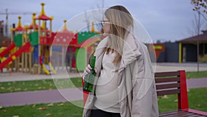Portrait of sad pregnant woman with beer bottle standing on childrens playground looking around thinking. Caucasian