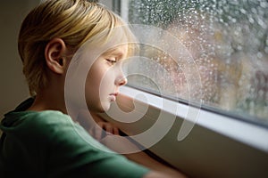 Portrait of a sad preteen boy standing at the window. Child watching the rain outside. Post-traumatic disorder. Accommodation of