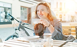 Portrait of sad red curled long hair caucasian teen girl sitting on a cozy cafe outdoor terrace on the street and drinking coffee