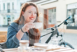 Portrait of sad red curled long hair caucasian teen girl sitting on a cozy cafe outdoor terrace on the street and drinking coffee