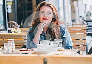Portrait of sad red curled long hair caucasian teen girl sitting on a cozy cafe outdoor terrace on the street and looking at the