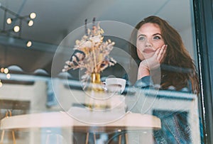 Portrait of sad red curled long hair caucasian teen girl sitting at table in cozy cafe and looking through the window glass at the