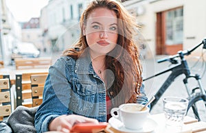 Portrait of sad red curled long hair caucasian woman sitting on a cozy cafe outdoor terrace on the street and looking at the