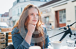 Portrait of sad red curled long hair caucasian woman sitting on a cozy outdoor cafe terrace on the street and looking at the
