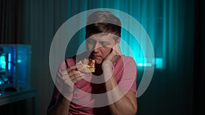 Portrait of sad tired young man eating pizza sitting alone at table in evening or night on blurred background of soft