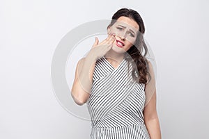 Portrait of sad unhappy beautiful young brunette woman with makeup and striped dress standing touching her chik because feel pain