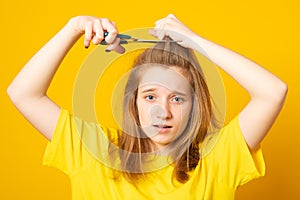 Portrait of sad and unhappy teen girl cutting her hair with scissors while standing over yellow background.