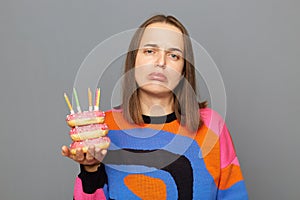Portrait of sad unhappy young adult woman with wearing warm sweater holding donut with candles isolated over gray background,
