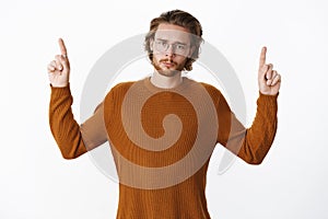 Portrait of sad unlucky and gloomy cute guy in glasses with beard making unhappy face as raising hands and pointing up