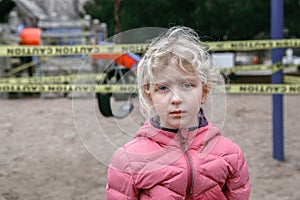 Portrait of sad upset Caucasian girl on closed playground outdoor. Kids play area locked with yellow caution tape in Toronto city