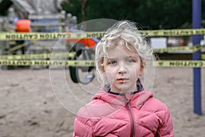 Portrait of sad upset Caucasian girl on closed playground outdoor. Kids play area locked with yellow caution tape in Toronto city