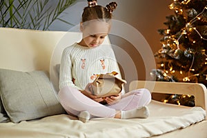 Portrait of sad upset little girl with two hair buns wearing casual clothing sitting on sofa with crossed legs, opens her