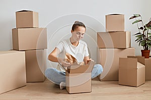 Portrait of sad upset woman wearing white T-shirt sitting on the floor near cardboard boxes, opening box with her belongings and