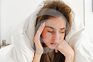 Portrait of sad woman feeling unwell, lying in bed under white blanket, touching head, has headache after hangover, has
