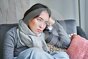 Portrait of sad woman feeling unwell, wearing warm clothes and scarf at her home without heating, staying on sick leave