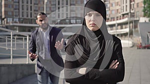 Portrait of sad woman in hijab standing with hands crossed and looking at camera as angry man yelling at the background