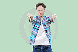 Portrait of sad young man in casual blue checkered shirt and headband standing, thumbs down and looking at camera with dislike and