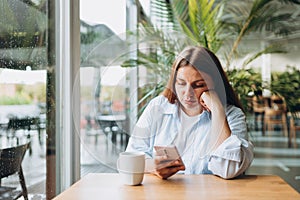 Portrait of sad young redhead woman in a coffee shop looking at smart phone. A lonely unhappy 30s girl siting in a cafe