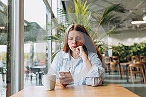 Portrait of sad young redhead woman in a coffee shop looking at smart phone. A lonely unhappy 30s girl siting in a cafe