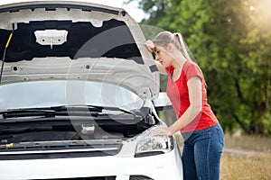 Portrait of sad young woman leaning on open car hood and looking at broken engine