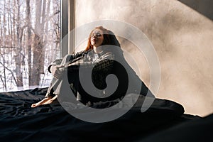 Portrait of sad young woman sitting by window on sunny day on windowsill with winter nature background, feels upset