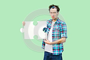 Portrait of sadness young adult man in checkered shirt standing and holding large piece of puzzle, thinking about solution of his