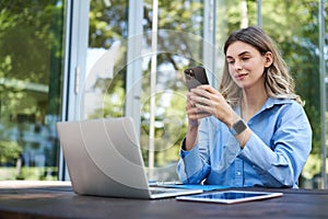 Portrait of saleswoman working outside office, sititng with laptop, using mobile phone. Digital nomad woman studying