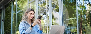 Portrait of saleswoman working outside office, sititng with laptop, using mobile phone. Digital nomad woman studying