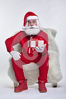 Portrait of a Santa Claus in sportsware sitting in a chair covered with sheepskin with gift box in hands