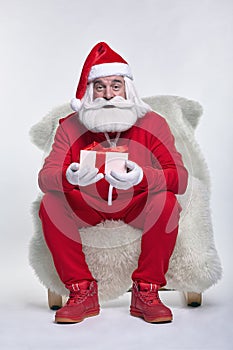 Portrait of a Santa Claus in sportsware sitting in a chair covered with sheepskin with gift box in hands