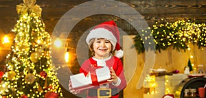 Portrait of Santa kid with gift near Christmas tree indoors. Christmas kids, happiness childhood concept.