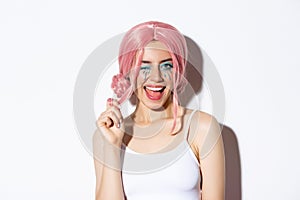 Portrait of sassy attractive girl in pink wig, wearing outfit for halloween party, smiling and winking at camera