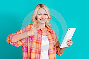 Portrait of satified woman with straight hair dressed plaid shirt holding tablet show thumb up isolated on turquoise