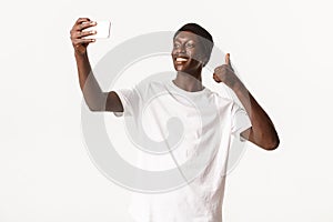 Portrait of satisfied african-american guy in beanie, smiling happy and showing thumbs-up while taking selfie or video