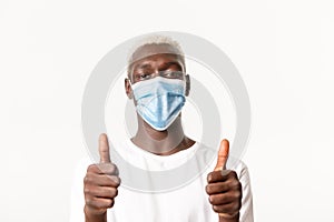 Portrait of satisfied african-american smiling blond guy, wearing medical mask and showing thumbs-up in approval, making