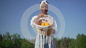 Portrait of satisfied African American woman showing basket with organic oranges looking at camera smiling. Happy