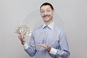 Portrait of satisfied attractive young adult businessman in blue shirt standing, giving and pointing with finger fan of money