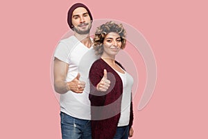 Portrait of satisfied couple of friends in casual style standing, hugging and showing thumbs up sign, looking at camera