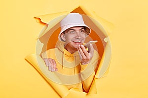 Portrait of satisfied delighted man wearing casual hoodie and panama looking through torn hole in yellow paper, talking to cell