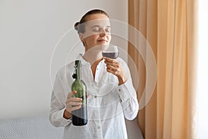 Portrait of satisfied delighted woman wearing white shirt holding bottle of alcoholic drink and smelling glass of wine, standing