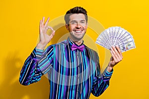 Portrait of satisfied guy dressed stylish shirt bow tie hold dollars show okey nice work isolated on vibrant yellow