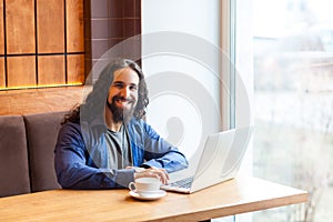 Portrait of satisfied handsome intelligence bearded young adult man freelancer in casual style sitting in cafe and working in