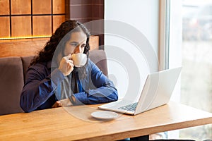 Portrait of satisfied handsome intelligence young adult man freelancer in casual style sitting in cafe with laptop, drinking cup