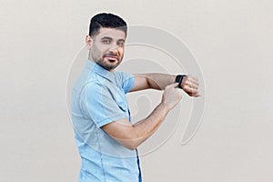 Portrait of satisfied handsome young bearded man in blue shirt standing, pointing and holding his smart watch, looking at camera