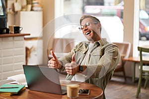 Portrait of satisfied, happy young man in glasses, working in cafe, sitting in co-working space with laptop, showing