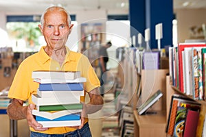 Portrait of satisfied older man in library with pile of books in hands