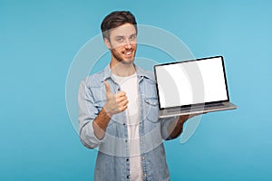 Portrait of satisfied smiling handsome man in worker denim shirt showing thumbs up and holding laptop